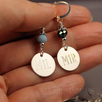 A hand holds two round silver stitch markers with beaded accents. Each has a disc engraved with “M1L” or “M1R.” The beads above the discs are light blue and faceted black. The background is soft and blurred. Text reads “LEILANI HANDMADE.”.