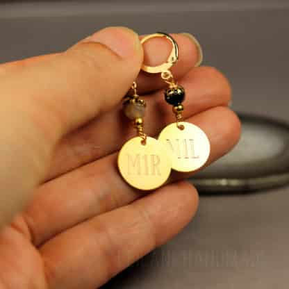 A hand holds two gold round charms engraved “M1R” and “M1L”. Each stitch marker features a small decorative bead above the charm. The background is blurred, highlighting the handmade jewelry details.