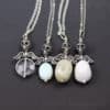 Four silver necklaces with winged skull pendants, each holding a different oval or round gemstone bead; beads are clear, light blue, pale green, and white. All are shown on a dark background labeled “leilani handmade.”.