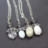 Four necklaces with silver skull and angel wing pendants, each holding a different oval gemstone: clear, light blue, light gray, and white. All are on silver chains, displayed on a dark surface. “Leilani handmade” is faintly visible at the bottom right.