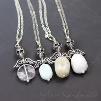 Four necklaces with silver skull and angel wing pendants, each holding a different oval gemstone: clear, light blue, light gray, and white. All are on silver chains, displayed on a dark surface. “Leilani handmade” is faintly visible at the bottom right.