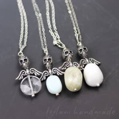 Four necklaces with silver skull and angel wing pendants, each holding a different oval gemstone: clear, light blue, light gray, and white. All are on silver chains, displayed on a dark surface. “Leilani handmade” is faintly visible at the bottom right.