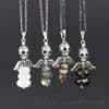Four necklaces with silver skull and wing pendants, each featuring three stacked gemstone beads in various colors—white, gray, green-pink, and dark brown—are displayed on silver chains against a dark background. The text “leilani handmade” appears below.