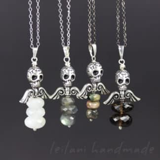Four necklaces with silver skull and wing pendants, each featuring three stacked gemstone beads in various colors—white, gray, green-pink, and dark brown—are displayed on silver chains against a dark background. The text “leilani handmade” appears below.