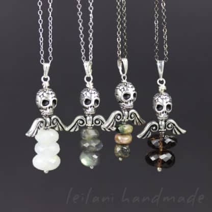 Four necklaces with silver skull and wing pendants, each featuring three stacked gemstone beads in various colors—white, gray, green-pink, and dark brown—are displayed on silver chains against a dark background. The text “leilani handmade” appears below.