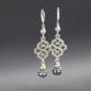 Two dangle celtic knot earrings with intricate silver loops, each featuring a white and a black pearl bead at the bottom, a small grey pearl with silver bead caps at the top. The earrings hang against a gray gradient background with faint "LEILANI HANDMADE" text at the bottom.