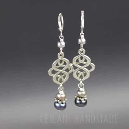 Two dangle celtic knot earrings with intricate silver loops, each featuring a white and a black pearl bead at the bottom, a small grey pearl with silver bead caps at the top. The earrings hang against a gray gradient background with faint "LEILANI HANDMADE" text at the bottom.