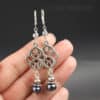 A hand holds two silver earrings with an intricate openwork knot pattern, each earring adorned with two beaded pearls—one light and one dark—below the knot design. The knot design is connected to the leverback earrings with a small grey pearl and smooth silver bead caps. Background is gray, with “LEILANI HANDMADE” faintly visible in the upper right.