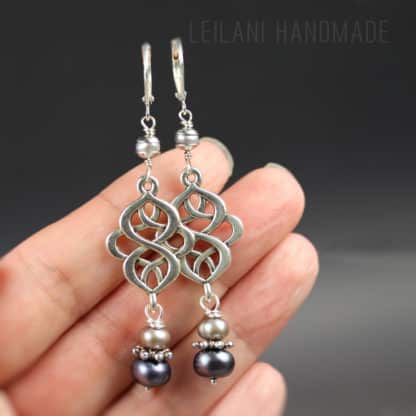 A hand holds two silver earrings with an intricate openwork knot pattern, each earring adorned with two beaded pearls—one light and one dark—below the knot design. The knot design is connected to the leverback earrings with a small grey pearl and smooth silver bead caps. Background is gray, with “LEILANI HANDMADE” faintly visible in the upper right.
