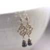 A pair of ornate silver dangle earrings hang on a ceramic bowl. Each earring features an intricate openwork design and three small pearl beads, one gray and one white, and a larger dark colored pearl at the bottom, separated by decorative silver spacers.
