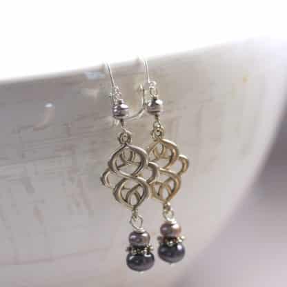 A pair of ornate silver dangle earrings hang on a ceramic bowl. Each earring features an intricate openwork design and three small pearl beads, one gray and one white, and a larger dark colored pearl at the bottom, separated by decorative silver spacers.