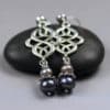 Two dangling earrings with intricate silver loops and dark and light pearl beads, resting on a smooth black stone against a gray background. Text “LEILANI HANDMADE” is faintly visible at the bottom of the image.
