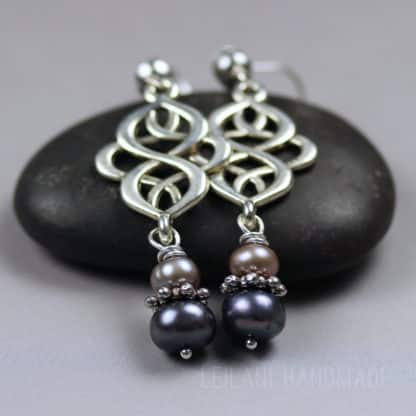 Two dangling earrings with intricate silver loops and dark and light pearl beads, resting on a smooth black stone against a gray background. Text “LEILANI HANDMADE” is faintly visible at the bottom of the image.