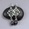 Two silver earrings rest on a smooth black stone. Each earring has an intricate, interwoven silver design and three pearls—one light, one medium, and one dark—separated by small, textured silver disks. Background is soft gray.