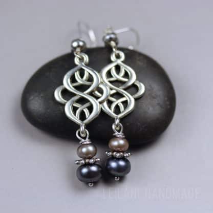 Two silver earrings rest on a smooth black stone. Each earring has an intricate, interwoven silver design and three pearls—one light, one medium, and one dark—separated by small, textured silver disks. Background is soft gray.