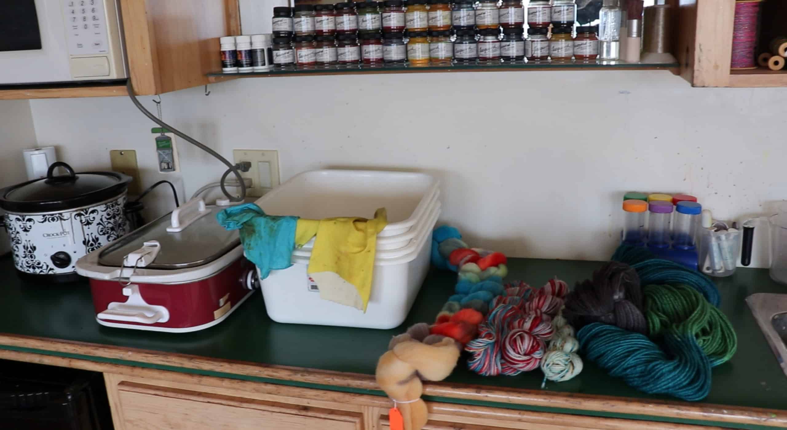 Skeins of colorful yarn and gloves sit on a green counter with two white tubs, dye bottles, a red slow cooker, and shelves of dye jars above. The scene appears to be set up for yarn dyeing.