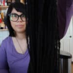 A person with glasses and straight dark hair, wearing a purple top, holds up a bundle of long black yarn or braids in front of their face, partially obscuring it, in a room with wooden shelves and books in the background.