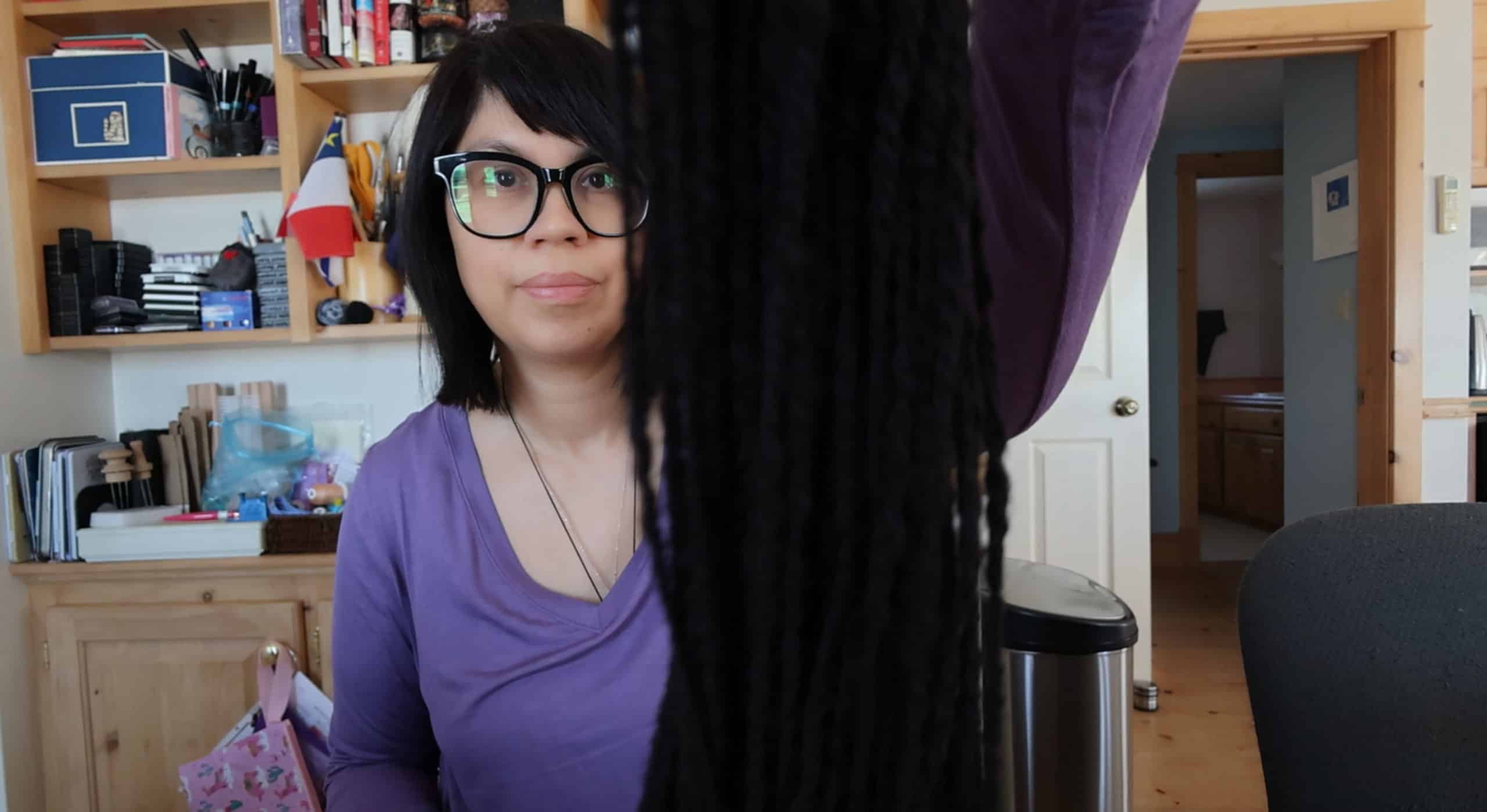 A person with glasses and straight dark hair, wearing a purple top, holds up a bundle of long black yarn or braids in front of their face, partially obscuring it, in a room with wooden shelves and books in the background.