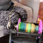 A person in patterned leggings sits on a couch, using a spinning wheel to ply targhee roving into colorful yarn onto a bobbin. The yarn is green, yellow, and blue. Hardwood floors and part of a sofa are visible in the background.