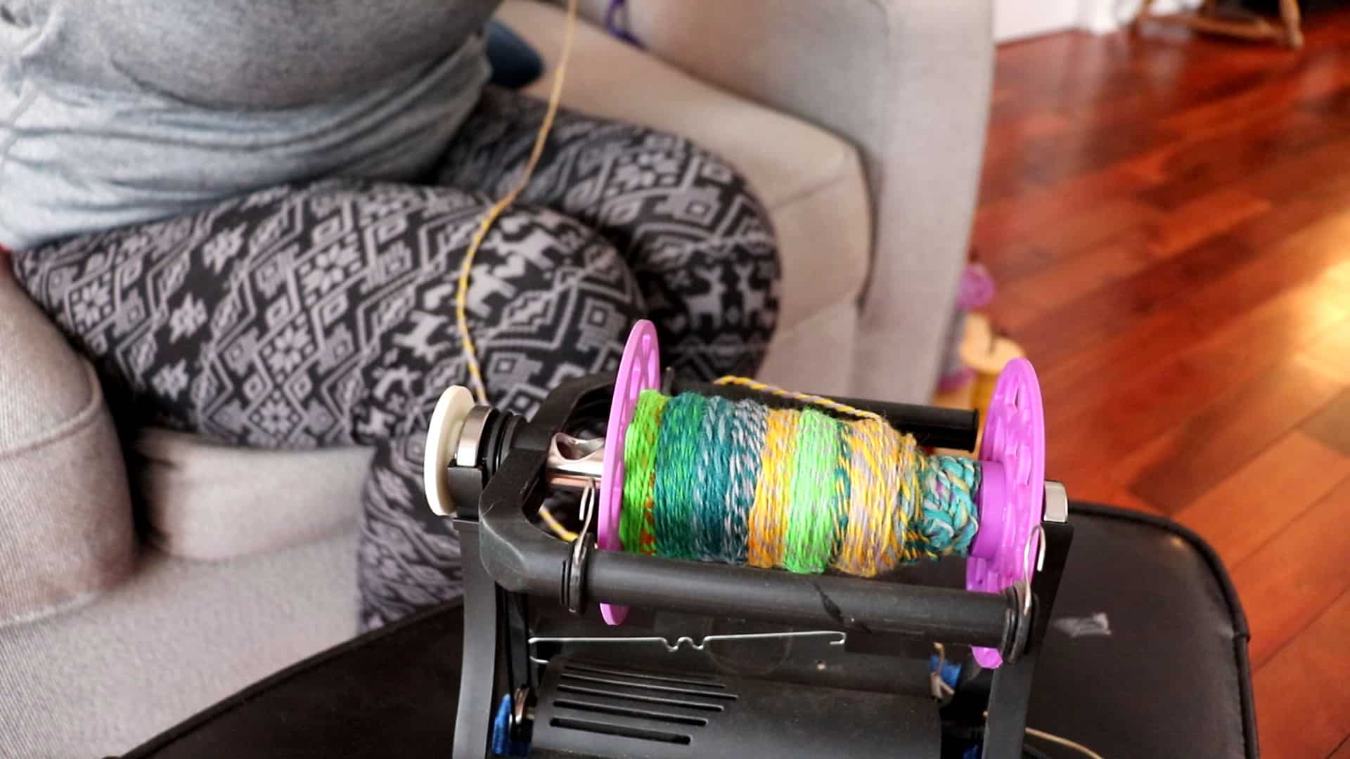 A person in patterned leggings sits on a couch, using a spinning wheel to ply targhee roving into colorful yarn onto a bobbin. The yarn is green, yellow, and blue. Hardwood floors and part of a sofa are visible in the background.