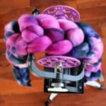 Overhead view of a spinning wheel with thick, braided, multicolored wool roving in shades of pink, purple, and blue draped over it, set on a tripod against a wooden floor background.