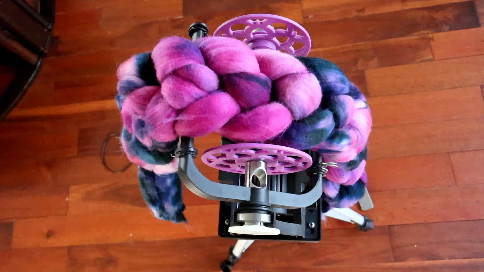 Overhead view of a spinning wheel with thick, braided, multicolored wool roving in shades of pink, purple, and blue draped over it, set on a tripod against a wooden floor background.